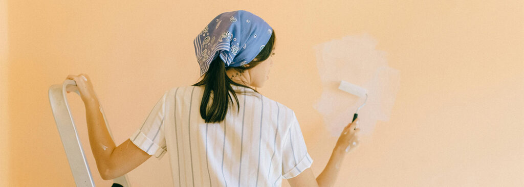 Woman in striped shirt and blue bandana painting over a peach wall using a small roller.

