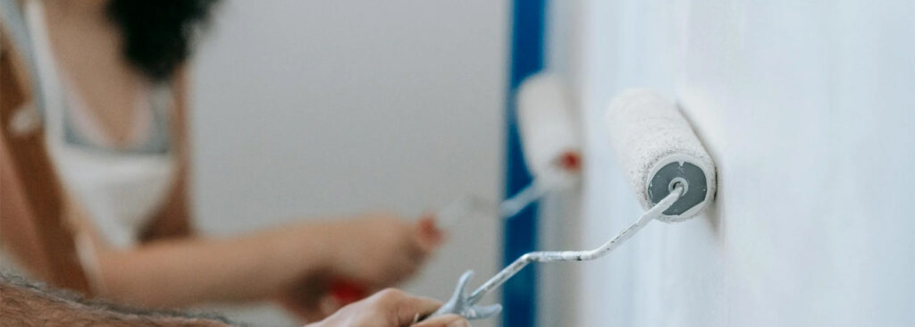 Two people using paint rollers to apply white primer to a wall.