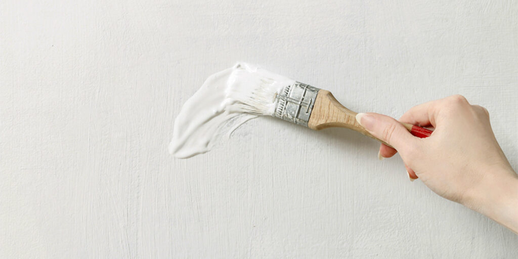Hand brushing white paint across a textured white wall.