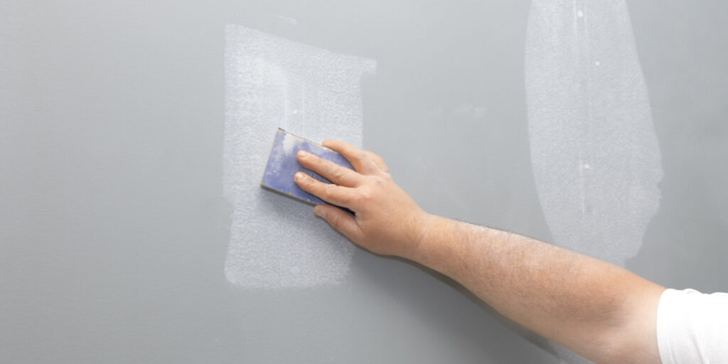 Hand sanding down a patched area on a grey wall.